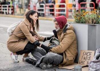 A young woman giving money to homeless beggar man sitting outdoors in city.