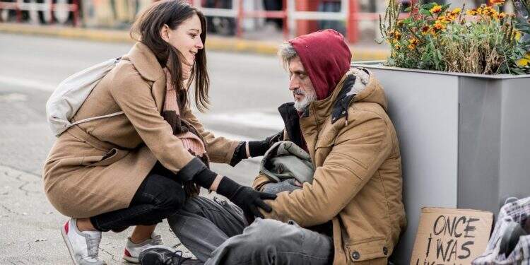 A young woman giving money to homeless beggar man sitting outdoors in city.